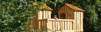 Zu sehen ist ein Baumhaus aus Holz. Es steht auf Stelzen. Über die Brüstung guckt ein dunkelhaariges Mädchen.