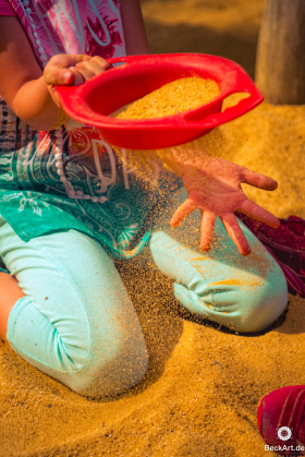 Kind (im Bils sichtbar bis zum Hals) sitzt im Sand und siebt ihn mit einem roten Sieb.