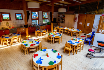 Speiseraum eines Kindergartens mit vielen Tischen und Stühlen. Die Tische sind mit buntem Geschirr gedeckt.